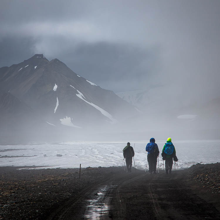 Trois marcheurs sur une piste noyée dans la brume, montagne sombre au fond