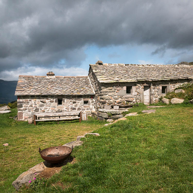 Les deux corps de bâtiment du buron et leur toit de lauzes, sous un ciel d'orage