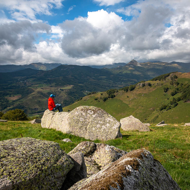 Randonneur assis sur un bloc de granit, face à la vallée et aux monts du Cantal